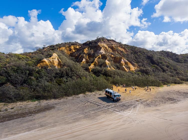 A tour vehicle parked on a beach as a group of people walk on the sand next to it