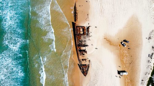 An aerial shot of a shipwreck on a beach with some tour vehicles parked near it